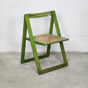 May include: A green folding chair with a woven seat. The chair has a simple design with a rectangular frame and a curved backrest. The chair is painted in a bright green color.