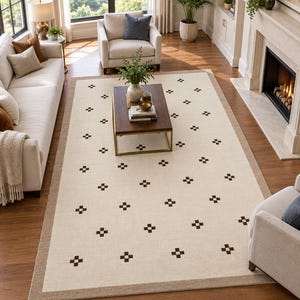 Puede incluir: Una alfombra rectangular que presenta un campo central de color crema con un patrón repetitivo de pequeñas cruces geométricas de color marrón oscuro. La alfombra está enmarcada por un borde ancho y sólido de color tostado en sus cuatro lados.
