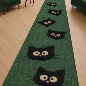 May include: A green runner rug featuring a repeating pattern of black cat faces with cream-colored eyes. The rug is placed on a light-colored wooden floor, with green armchairs and a side table in the background.