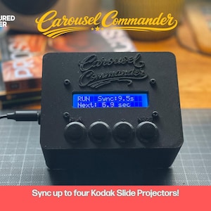 Op de afbeelding: Zwart elektronisch apparaat met de tekst "Carousel Commander" en een blauw scherm met "RUN Sync: 9.5s Next: 6.9 sec." Het apparaat heeft vier zwarte knoppen en een kabel. De tekst "Sync up to four Kodak Slide Projectors!" staat onderaan.