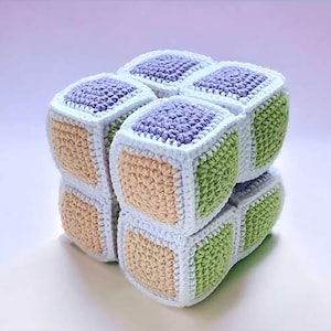 Könnte beinhalten: Ein weiches Häkel-Puzzle-Würfelspielzeug, das aus acht kleineren, miteinander verbundenen Einzelwürfeln besteht. Jede Seite zeigt ein strukturiertes Quadrat in Lavendel, Hellgrün oder Beige, umrandet von dickem weißem Garn.
