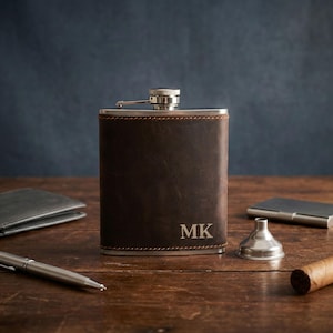 May include: A brown leather flask with the initials "MK" engraved on the front. The flask is stainless steel and has a silver cap. Other items on the wooden surface include a wallet, pen, business card holder, funnel, and cigar.