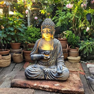 May include: A weathered-look Buddha statue in a seated meditation pose. The statue is gray and brown with a warm, glowing light emanating from within. It sits on a wooden base, surrounded by lush greenery, creating a serene and spiritual ambiance.