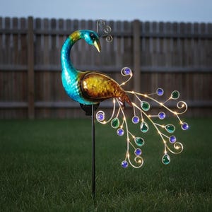 May include: A colorful peacock garden stake with a blue, yellow, and bronze body. The peacock's tail features blue and green jewel-like accents with warm white lights. The stake is made of metal and is placed in a grassy yard.