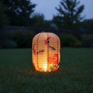 May include: A glowing, paper lantern with a floral and fish design, illuminated from within. The lantern is cylindrical with a rounded top and bottom, and is set on a grassy lawn. The background features a blurred view of trees and foliage.