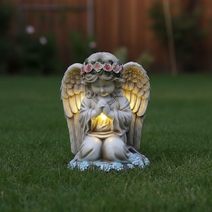 May include: A kneeling angel statue with a floral crown and illuminated wings and hands. The statue is beige and sits on a bed of blue and white flowers, placed on a green lawn. The angel's wings are lit from behind.