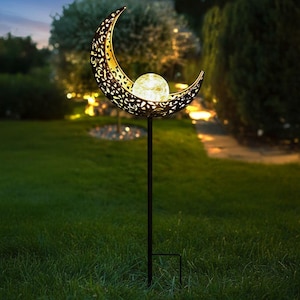 May include: A decorative outdoor solar garden light featuring a crescent moon design with intricate cut-out patterns. A glowing orb sits within the moon's embrace, mounted on a black metal stake for easy placement in a garden or yard.