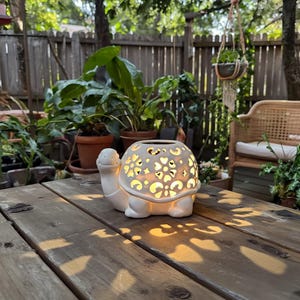 Puede incluir: Un portavelas de cerámica blanca con forma de tortuga y recortes en forma de corazón. El portavelas está sobre una mesa de madera. El portavelas está encendido, proyectando un brillo cálido y sombras en forma de corazón.