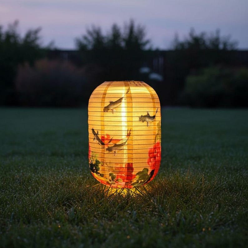 May include: A glowing, paper lantern with a fish and floral design, illuminated from within. The cylindrical lantern is yellow with black horizontal stripes and stands on a grassy lawn. The design includes red flowers and gray fish.