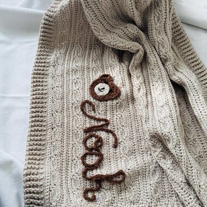 Op de afbeelding: Een beige kabelgebreide babydeken met een bruine berenapplicatie en de naam "Noah" in bruin cursief geborduurd. De deken heeft een textuurpatroon en een gehaakte rand.