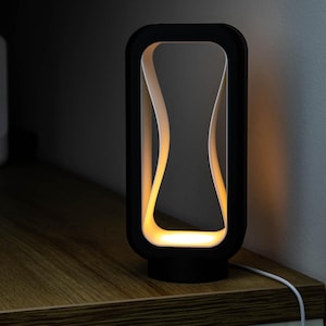 May include: A modern black table lamp with a unique hourglass-shaped light design. The lamp has a rectangular frame with rounded corners and a warm, yellow glow emanating from within. It sits on a wooden surface, with a white cord extending from the base.
