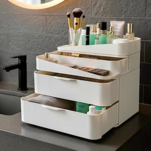 May include: A white, tiered makeup organizer with three pull-out drawers. The top tier holds makeup brushes and bottles. The drawers contain makeup, a towel, and skincare products. The organizer sits on a gray countertop.