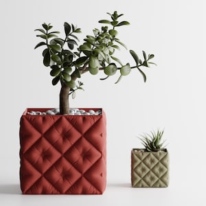 Op de afbeelding: Twee decoratieve vierkante plantenbakken, één groot en één klein, met een gewatteerd ontwerp. De grotere plantenbak is dieprood en bevat een jadeplant. De kleinere plantenbak is olijfgroen en bevat een kleine vetplant.
