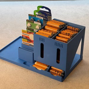 May include: A blue battery organizer with multiple compartments. It holds various sizes of batteries, including AA and button cell batteries. The organizer is designed to keep batteries neatly stored and accessible.