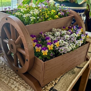 May include: A brown wooden wagon planter filled with colourful flowers. The planter has two levels and decorative wheels. The flowers include white, yellow, and purple blooms, creating a vibrant display for a garden or patio.