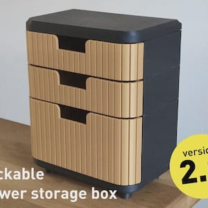 Könnte beinhalten: Eine stapelbare Schubladen-Aufbewahrungsbox mit drei Schubladen. Die Box ist überwiegend dunkelgrau mit hellbraunen Schubladenfronten. Die Schubladenfronten haben ein vertikales Rippendesign. Der Text "Version 2.0" befindet sich auf einem gelben Kreis.
