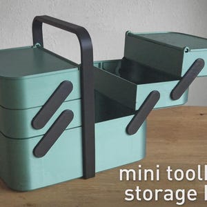 Può includere: Una mini cassetta degli attrezzi verde menta con manico nero e chiusure nere. La scatola ha più scomparti ed è aperta, rivelando l'interno. Il testo "mini toolbox storage box" è visualizzato in bianco.