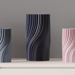 May include: Three cylindrical vases in shades of gray, black, and pink. Each vase features a vertical, wave-like design. The vases are displayed on a white surface against a gray background. The tallest vase is approximately 10 inches tall.
