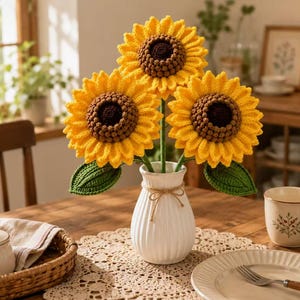 Op de afbeelding: Drie gehaakte zonnebloemen met felgele bloemblaadjes en donkerbruine harten, elk op een groene steel met groene bladeren, geschikt in een witte keramische vaas die is vastgebonden met een dunne strik van touw. Zichtbare textuur van garen.