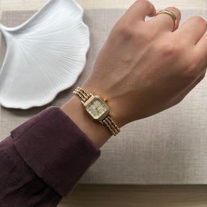 Könnte beinhalten: Eine goldfarbene Armbanduhr mit quadratischem Zifferblatt und Gliederarmband. Die Uhr hat ein helles Zifferblatt mit schwarzen Zeigern und Markierungen. Das Armband besteht aus kleinen, verbundenen Gliedern. Ein goldener Ring wird am Finger getragen.