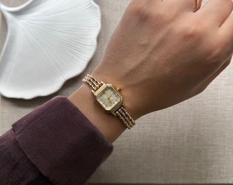 Puede incluir: Un reloj de pulsera dorado con una esfera cuadrada y una correa de eslabones. El reloj tiene una esfera de color claro con manecillas y marcadores negros. La correa está hecha de pequeños eslabones conectados. Un anillo dorado se lleva en un dedo.