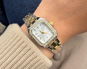 May include: A rectangular quartz wristwatch featuring a two-tone silver and gold metal link band. The watch face has a white grid pattern, gold hands, and a bezel encrusted with small clear stones. A gold crown is on the side of the case.
