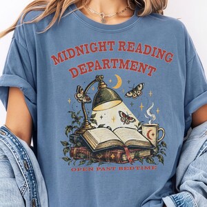 以下が含まれることがあります： MIDNIGHT READING DEPARTMENTおよびOPEN PAST BEDTIMEのテキストがプリントされた青いTシャツ。デザインには、開いた本、点灯したデスクランプ、蛾、三日月、月アイコン付きの湯気の立つマグカップが描かれています。