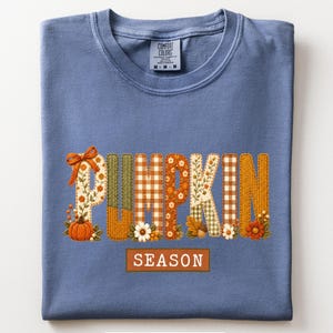 May include: A blue jean colored t-shirt featuring the word PUMPKIN in large, patterned letters with floral, plaid, and knit textures, accented by a pumpkin and flowers. Below, a rectangular orange box contains the white text SEASON.