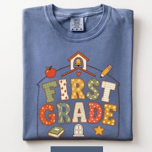 May include: A blue jean colored t-shirt featuring a schoolhouse outline graphic with the text "FIRST GRADE" in patterned block letters. The design includes icons of an apple, a bell, a pencil, a book, and a star, all printed on the front of the shirt.