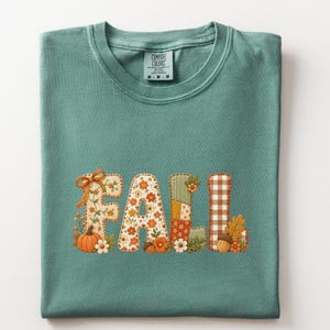 May include: A light green t-shirt featuring the word FALL in large letters. Each letter has a unique pattern including floral prints, brown and white gingham, and solid patches. The design includes a small orange pumpkin and an acorn.