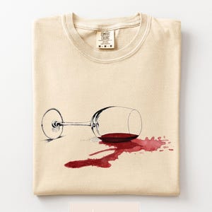 May include: Ivory-colored t-shirt featuring a graphic of a tipped-over wine glass with spilled red wine. The Comfort Colors brand label is visible at the top. The shirt is folded neatly.