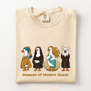 May include: An ivory t-shirt featuring a graphic of four ducks dressed as famous art subjects, including a pearl earring, Mona Lisa, Venus, and The Scream. The text "Museum of Modern Quack" is printed below the ducks in a brown, sans-serif font.
