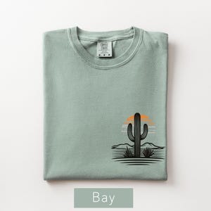 Puede incluir: Una camiseta de manga corta color verde salvia con un gr&aacute;fico negro de un cactus saguaro en un paisaje des&eacute;rtico con un sol naranja. La prenda tiene cuello redondo y una etiqueta de la marca Comfort Colors impresa en el interior del cuello.