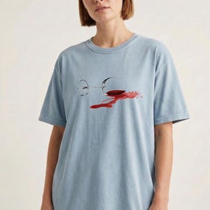 May include: A light blue short-sleeve t-shirt featuring a graphic print of a tipped-over wine glass with a red wine spill. The design shows the glass lying on its side with a dark red liquid puddle spreading out from the base of the glass.