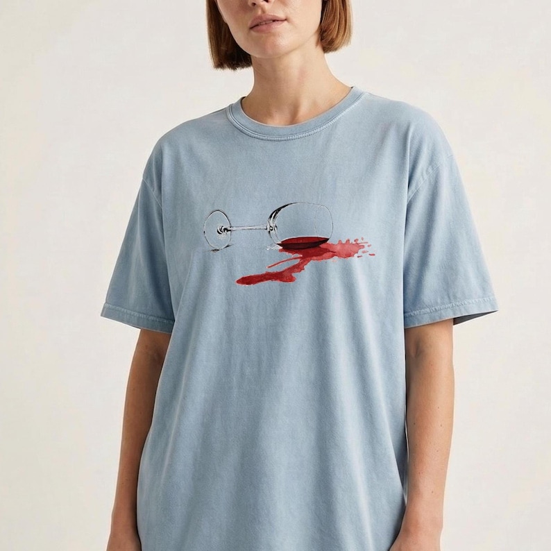 May include: A light blue short-sleeve t-shirt featuring a graphic print of a tipped-over wine glass with a red wine spill. The design shows the glass lying on its side with a dark red liquid puddle spreading out from the base of the glass.