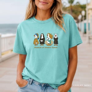 May include: A light mint green short-sleeve t-shirt featuring a graphic print of four ducks dressed as famous art figures. Below the ducks, the text reads Museum of Modern Quack. The shirt has a crew neckline and a relaxed, casual fit.