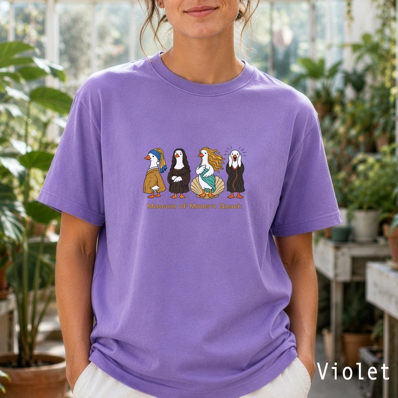 May include: A violet short-sleeved t-shirt featuring a graphic print of four ducks dressed as famous art figures. Below the ducks, the text reads "Museum of Modern Quack". The shirt has a crew neckline and a relaxed fit, showcasing a vibrant purple hue.