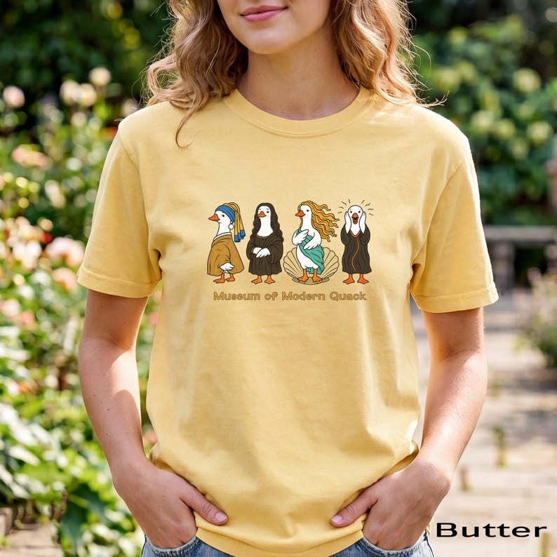 May include: A butter yellow t-shirt featuring a graphic of four ducks dressed as famous art figures. Below the ducks, the text reads "Museum of Modern Quack". The design includes a duck with a blue headscarf, a duck in a black robe, a duck on a shell, and a duck with hands on its face.