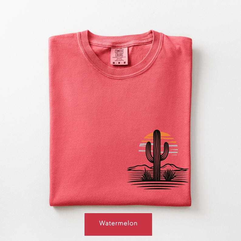 Puede incluir: Una camiseta color sand&iacute;a con un gr&aacute;fico negro de un cactus saguaro en un paisaje des&eacute;rtico con un sol amarillo y naranja detr&aacute;s. La etiqueta interior del cuello muestra el texto Comfort Colors y la talla M. La camiseta est&aacute; doblada.