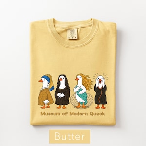 May include: A butter yellow t-shirt featuring a graphic of four ducks dressed as famous art figures. Below the ducks, the text reads Museum of Modern Quack. The shirt has a crew neckline and a Comfort Colors brand label visible at the inner collar.
