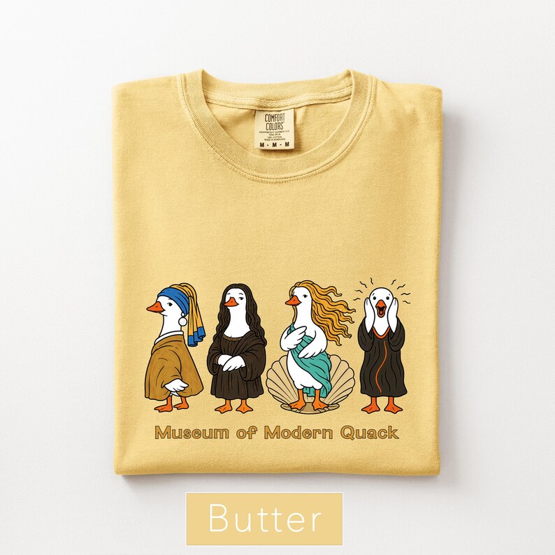 May include: A butter yellow t-shirt featuring a graphic of four ducks dressed as famous art figures. Below the ducks, the text reads Museum of Modern Quack. The shirt has a crew neckline and a Comfort Colors brand label visible at the inner collar.
