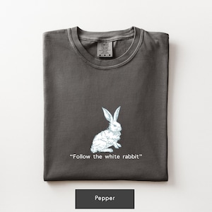 May include: A dark grey t-shirt with a white rabbit illustration and the text "Follow the white rabbit." The shirt is folded and has a crew neck. The bottom of the shirt has a black label that says "Pepper."