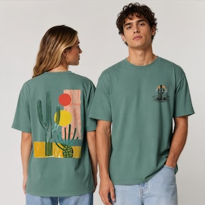 Puede incluir: Una camiseta de manga corta color verde salvia con un gr&aacute;fico en la espalda que muestra un cactus saguaro alto, un cactus redondo m&aacute;s peque&ntilde;o, un c&iacute;rculo rojo, un c&iacute;rculo amarillo y un bloque rectangular color melocot&oacute;n. El pecho muestra un icono de cactus.