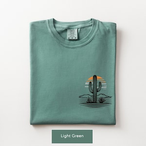 Puede incluir: Una camiseta de manga corta de color verde claro con un gr&aacute;fico de la silueta de un cactus negro sobre un sol a rayas naranja y blanco. La prenda tiene cuello redondo y una etiqueta de la marca Comfort Colors impresa en el interior.