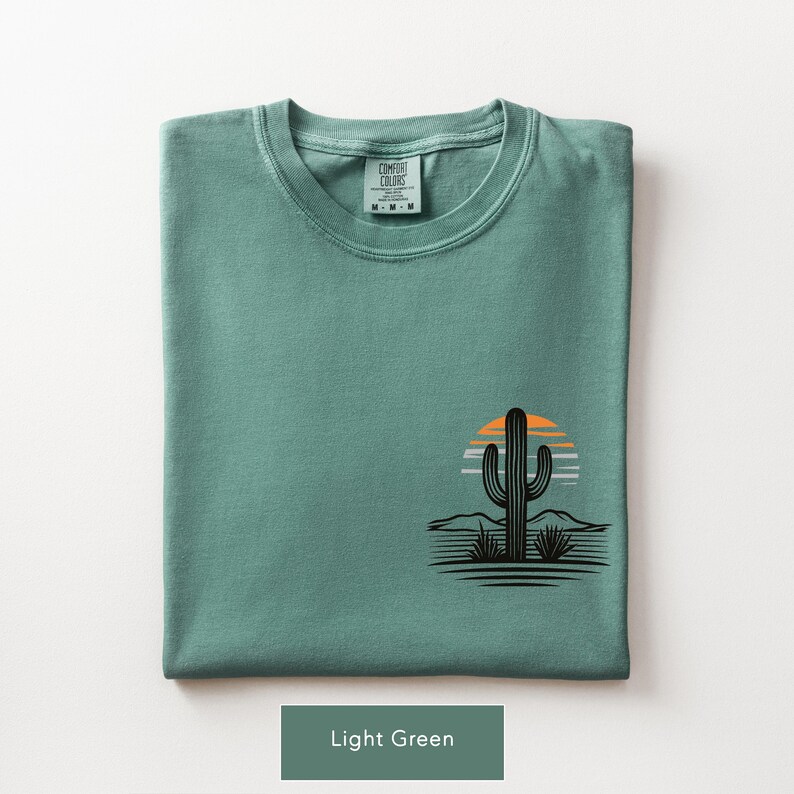 Puede incluir: Una camiseta de manga corta de color verde claro con un gr&aacute;fico de la silueta de un cactus negro sobre un sol a rayas naranja y blanco. La prenda tiene cuello redondo y una etiqueta de la marca Comfort Colors impresa en el interior.