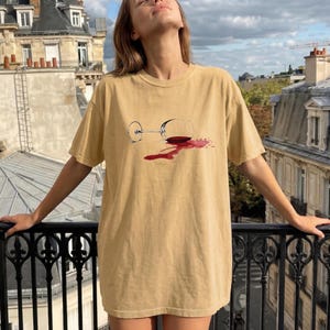 May include: A tan short-sleeved t-shirt featuring a graphic print of a clear wine glass lying on its side with a pool of spilled red wine spreading across the fabric. The shirt has a crew neckline and a relaxed, loose-fitting silhouette.