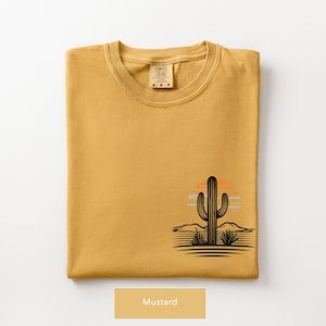 Puede incluir: Una camiseta de cuello redondo color amarillo mostaza con un gr&aacute;fico en la parte inferior derecha. El dise&ntilde;o muestra la silueta negra de un cactus saguaro en un paisaje des&eacute;rtico con un sol estilizado en naranja y azul claro tras las monta&ntilde;as.