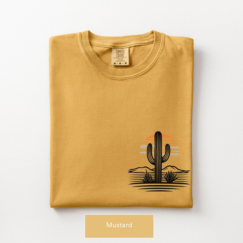 Puede incluir: Una camiseta de cuello redondo color amarillo mostaza con un gr&aacute;fico en la parte inferior derecha. El dise&ntilde;o muestra la silueta negra de un cactus saguaro en un paisaje des&eacute;rtico con un sol estilizado en naranja y azul claro tras las monta&ntilde;as.