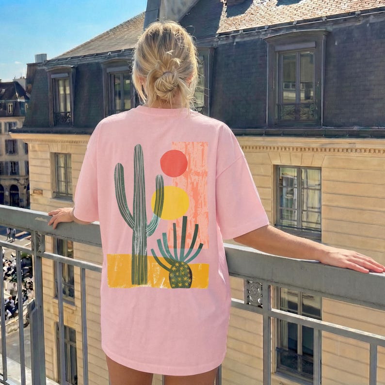 Puede incluir: Una camiseta de manga corta de color rosa claro con un estampado gr&aacute;fico en la espalda. El dise&ntilde;o muestra un cactus verde alto, un cactus redondo m&aacute;s peque&ntilde;o, un c&iacute;rculo rojo, un c&iacute;rculo amarillo y un bloque naranja texturizado.