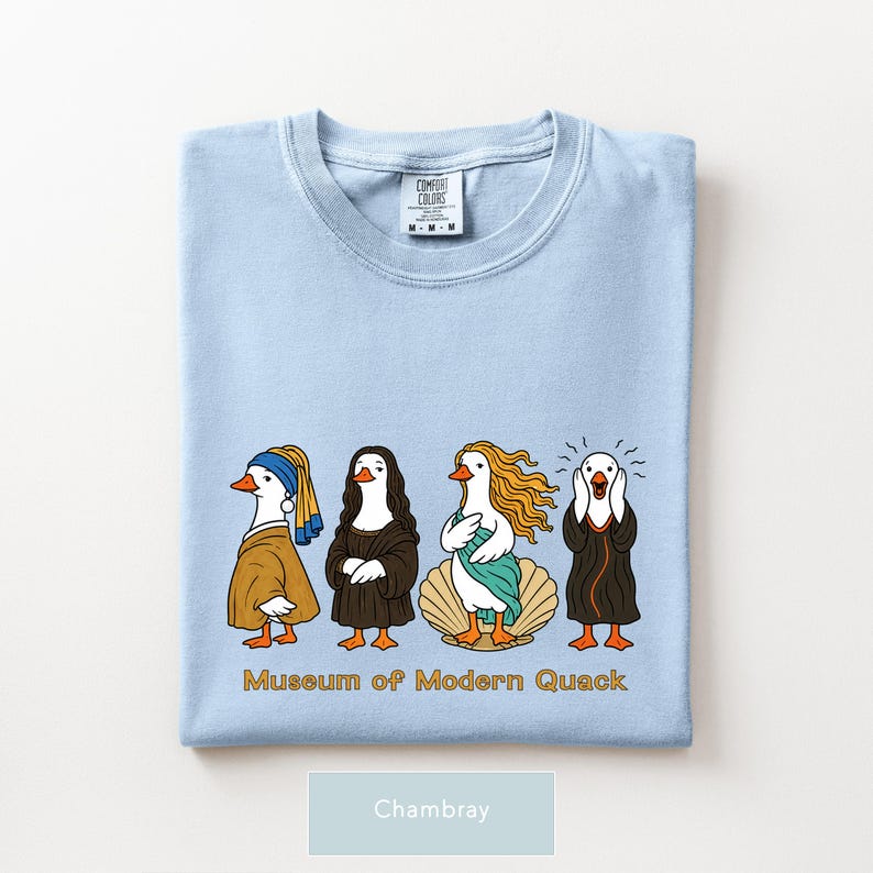 May include: A light blue t-shirt featuring a graphic of four ducks dressed as famous art subjects, including a pearl earring, Mona Lisa, Birth of Venus, and The Scream. The text "Museum of Modern Quack" is printed in gold below the ducks.
